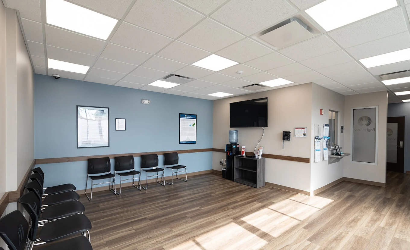 BrightView Health waiting area with rows of black chairs, blue accent wall, wall-mounted television, water dispenser, informational displays, and wood-look flooring