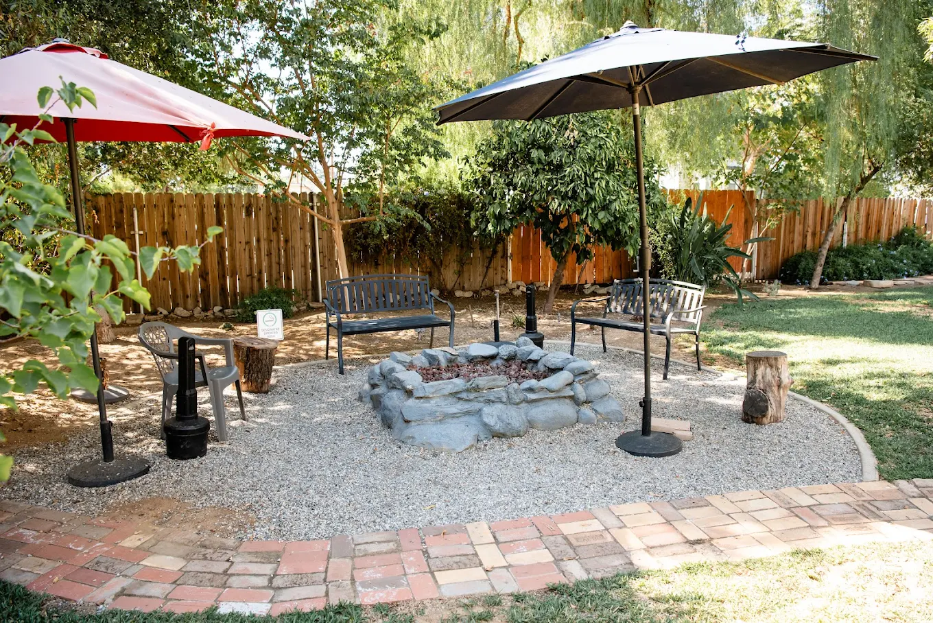 Firepit seating area with chairs and umbrellas on gravel surrounded by trees and fencing