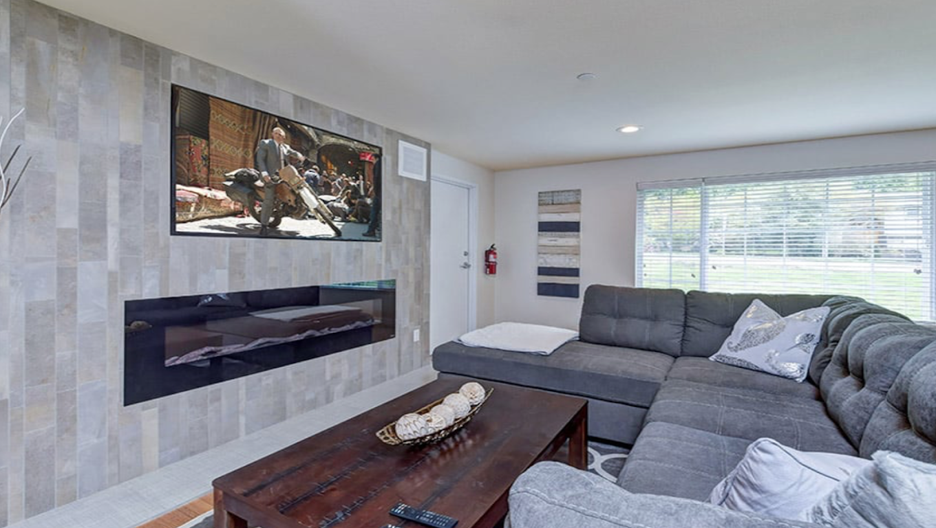 Cozy living space with sectional sofa, wall-mounted TV, and modern electric fireplace, illuminated by natural light from a large window
