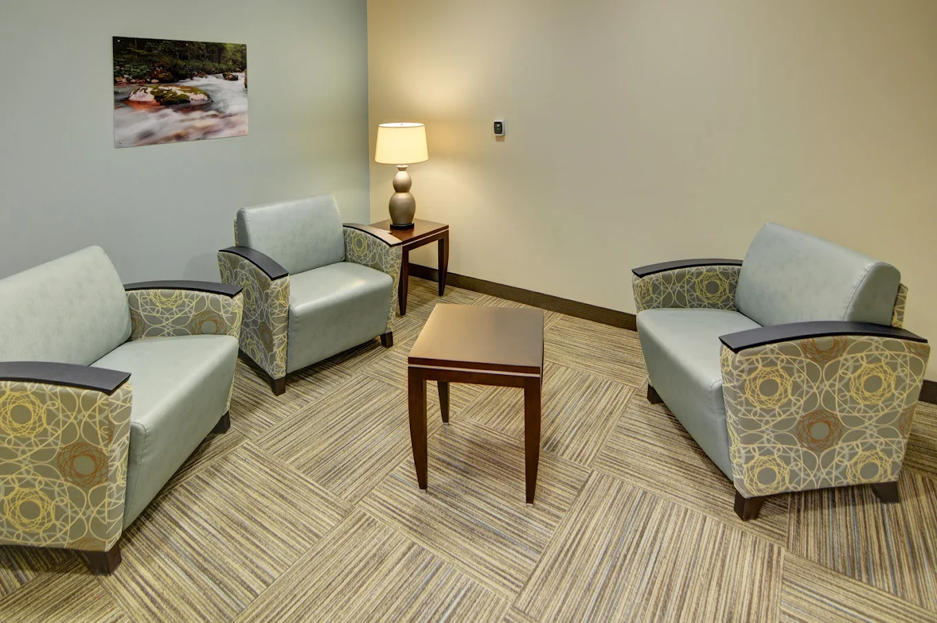 A quiet therapy room with four cushioned armchairs arranged around a small wooden table, softly lit by a lamp for a calm atmosphere.