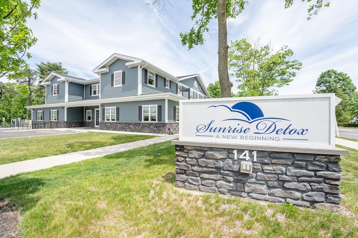  A sign in front of the facility reading "Sunrise Detox - A New Beginning," placed on a stone base with neatly landscaped greenery.