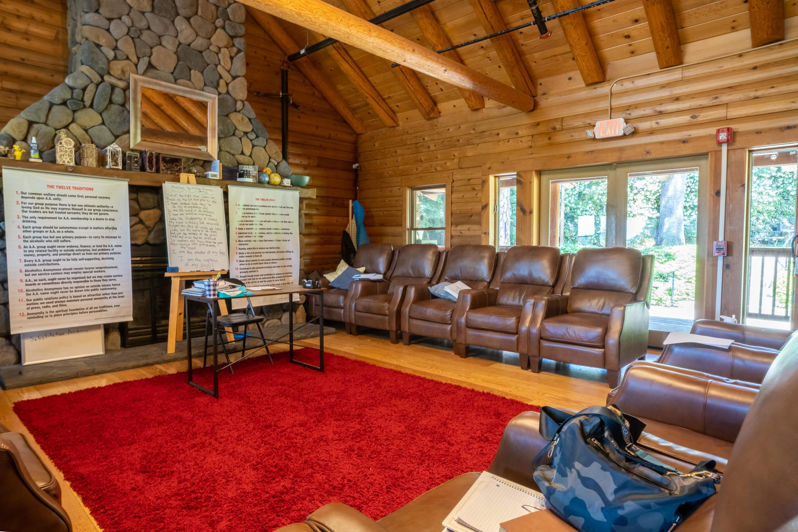 Warm, cabin-style therapy room with leather chairs arranged in a circle, stone fireplace, and AA recovery posters displayed