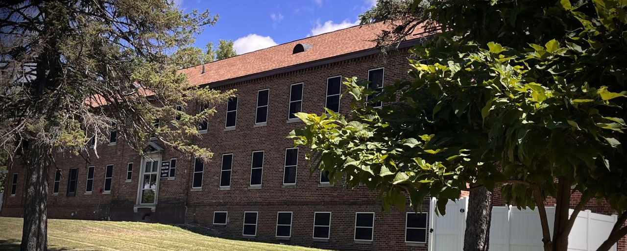  A large brick building with multiple windows and a sloped roof, surrounded by greenery.