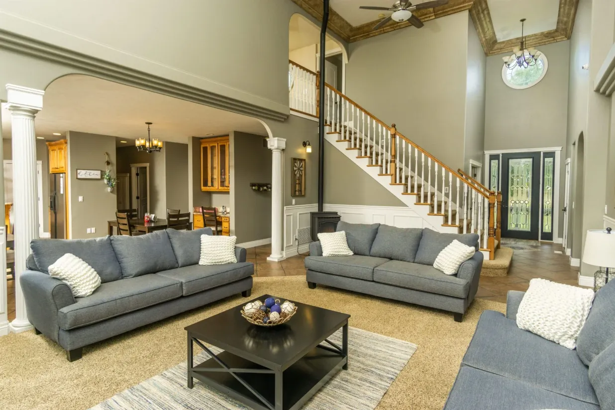 Spacious living room with gray sofas, high ceilings, staircase, and view into the kitchen and dining area