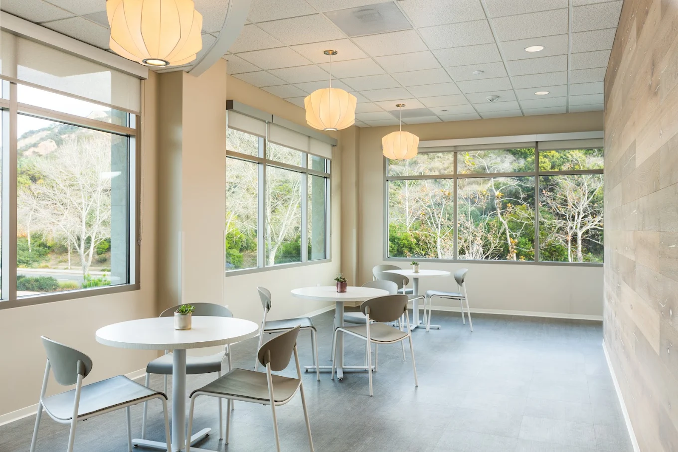 Bright recreational lounge with round tables and chairs, pendant lighting, and large windows overlooking trees