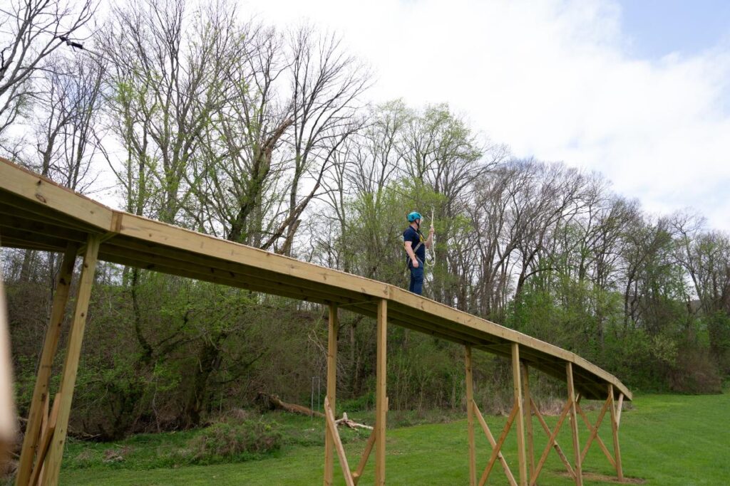 Person walking along an elevated wooden balance beam course in a harness, surrounded by trees