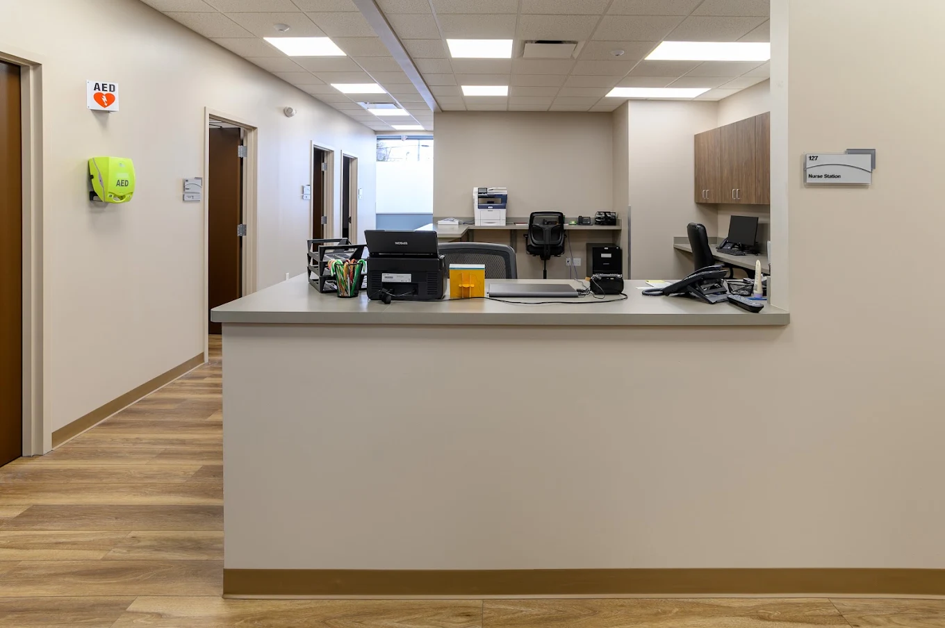 BrightView Health nurse station with reception counter, computers, phones, office workstations, wall cabinets, AED equipment, and clinical hallway