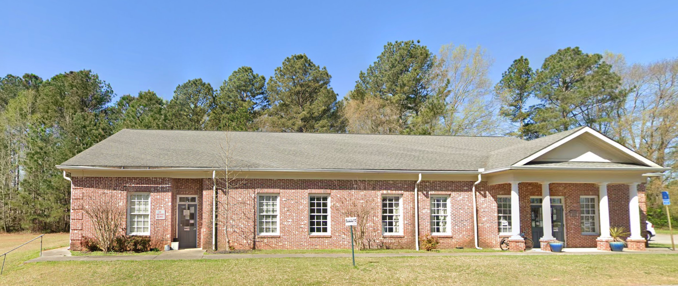 A single-story red brick building with white columns at the entrance, surrounded by green trees and a well-maintained lawn, serving as the Advantage Behavioral Health Systems Walton County Mental Health Center.