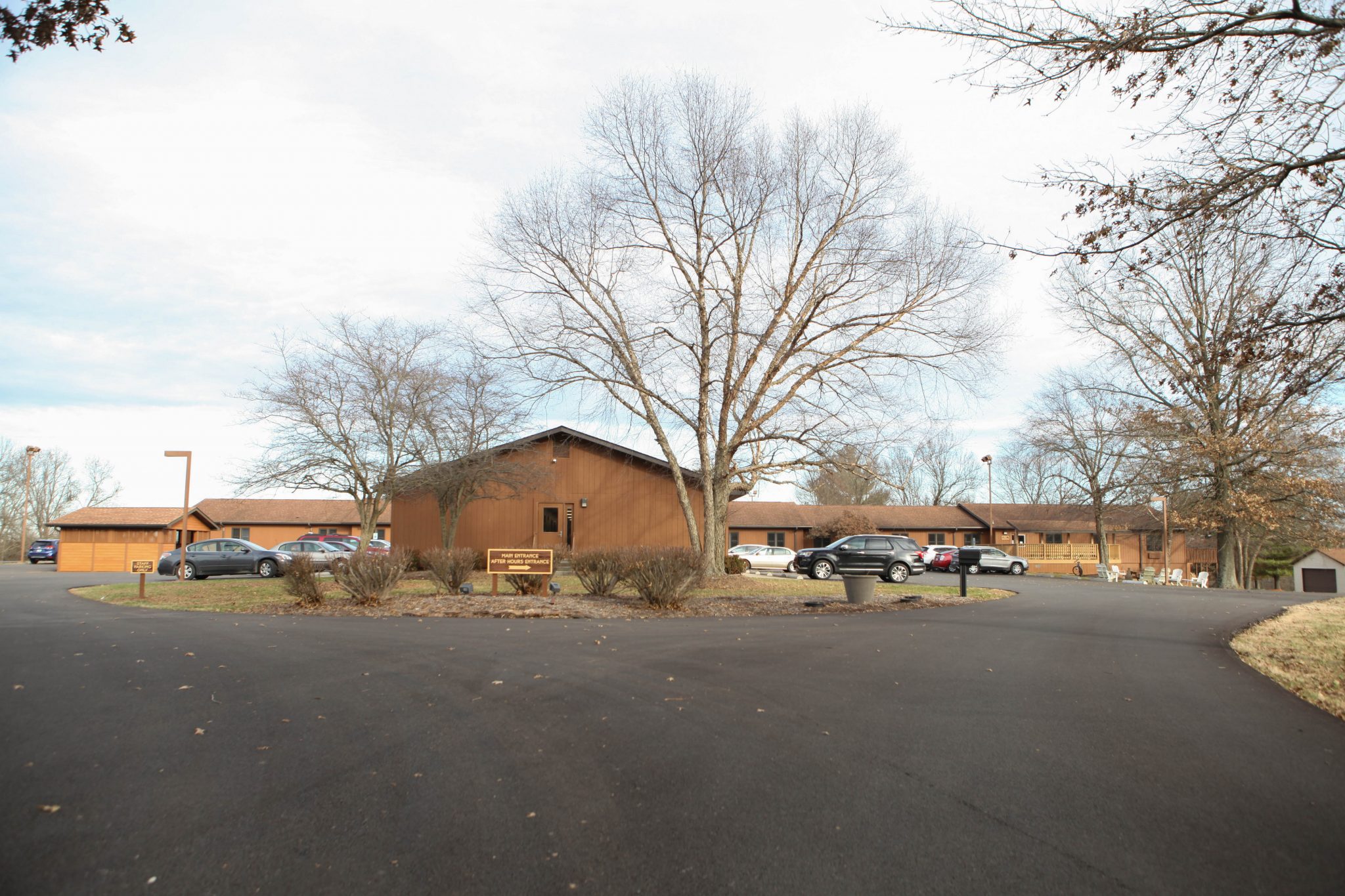 Brown buildings and parking area surrounded by leafless trees on a clear day.