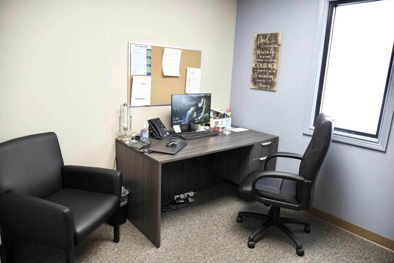 Private consultation office at BrightView Health with desk, computer, telephone, visitor chair, office chair, bulletin board, window, and blue accent wall