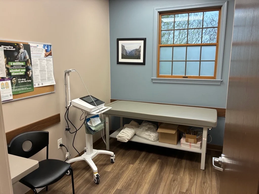 Medical exam room with examination table, clinical equipment, patient chair, bulletin board, window, and medical supplies