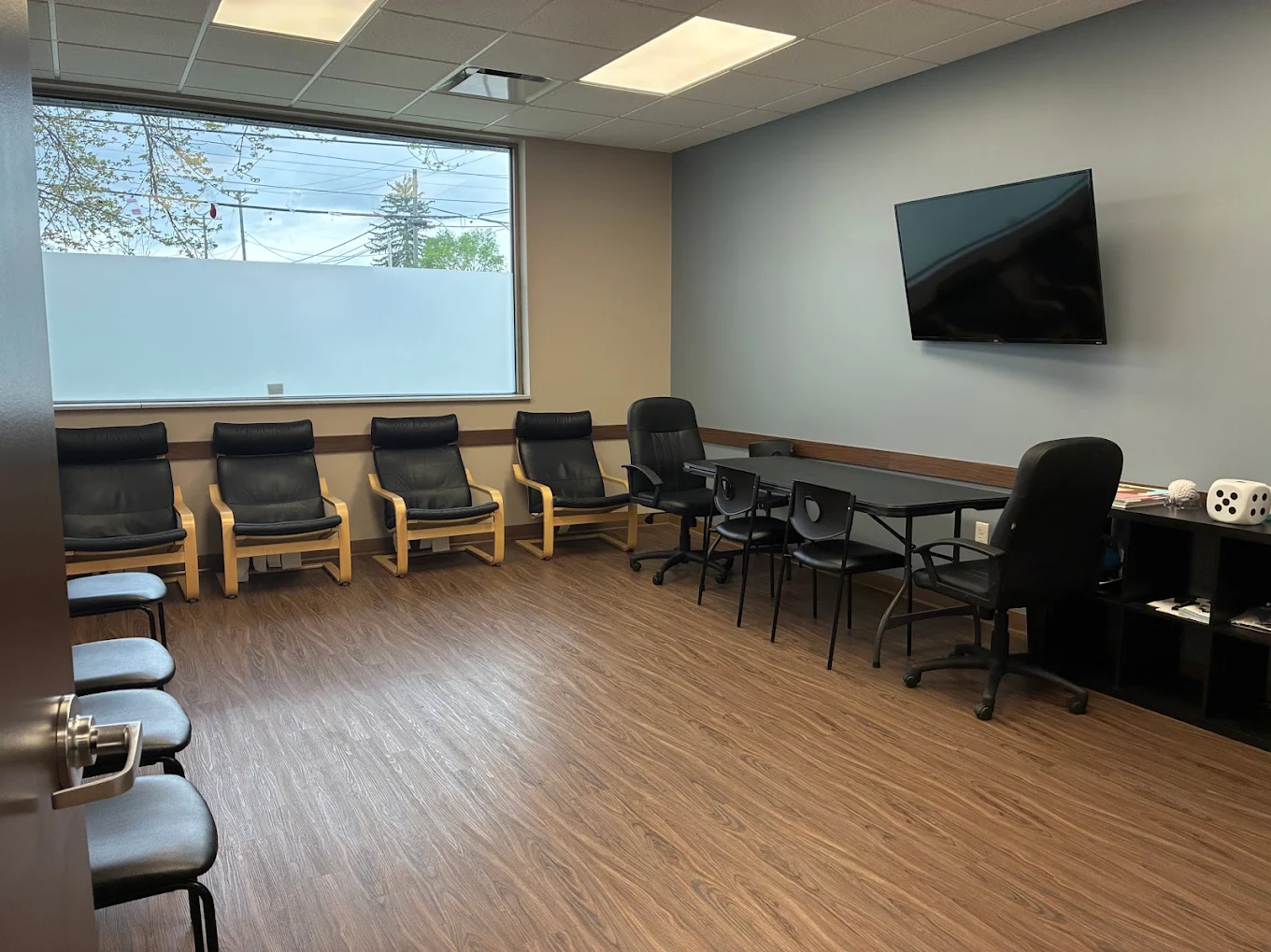 Group counseling area with multiple chairs, tables, wall-mounted television, large window, and storage shelving