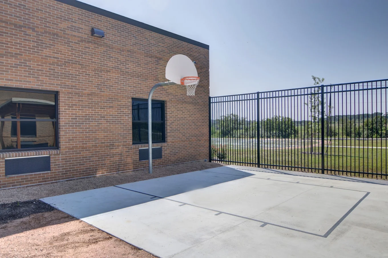 A fenced outdoor basketball half-court next to a brick building, featuring a single hoop and a concrete playing surface.