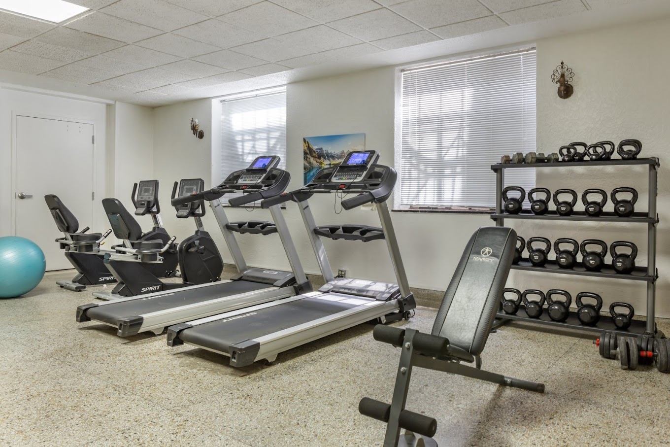 A small fitness room with treadmills, stationary bikes, weight racks, and a workout bench in a clean, well-lit space.