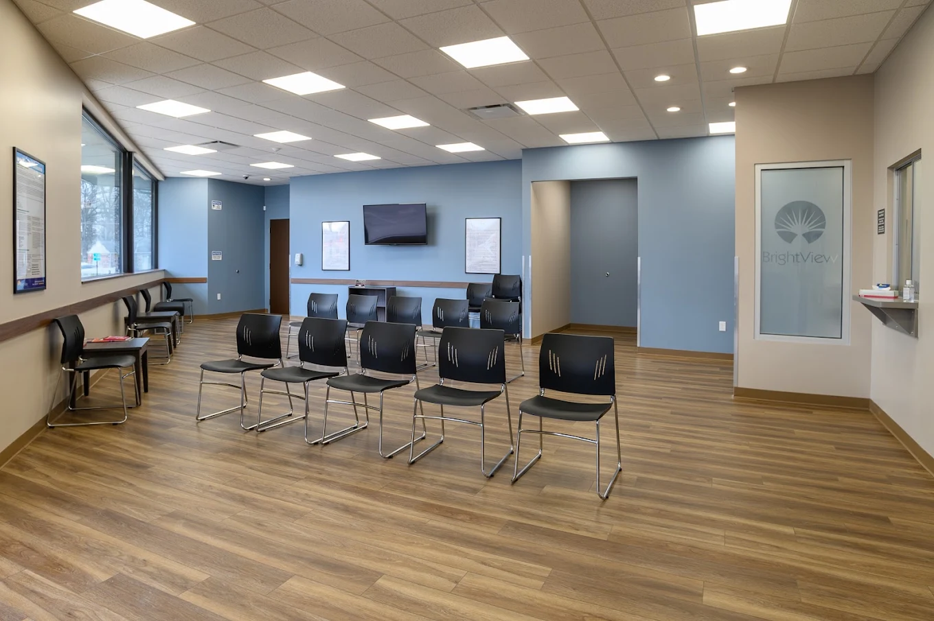 Spacious BrightView Health waiting area with rows of black chairs, wood-look flooring, blue accent wall, large windows, television, and service windows