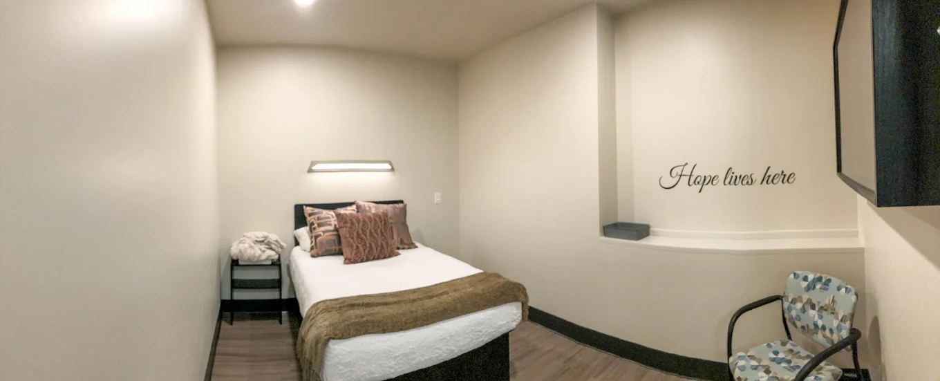 A cozy single bedroom with neutral-colored walls, a twin bed with accent pillows and throw, a small side table, and a chair by the wall with “Hope lives here” written above it.