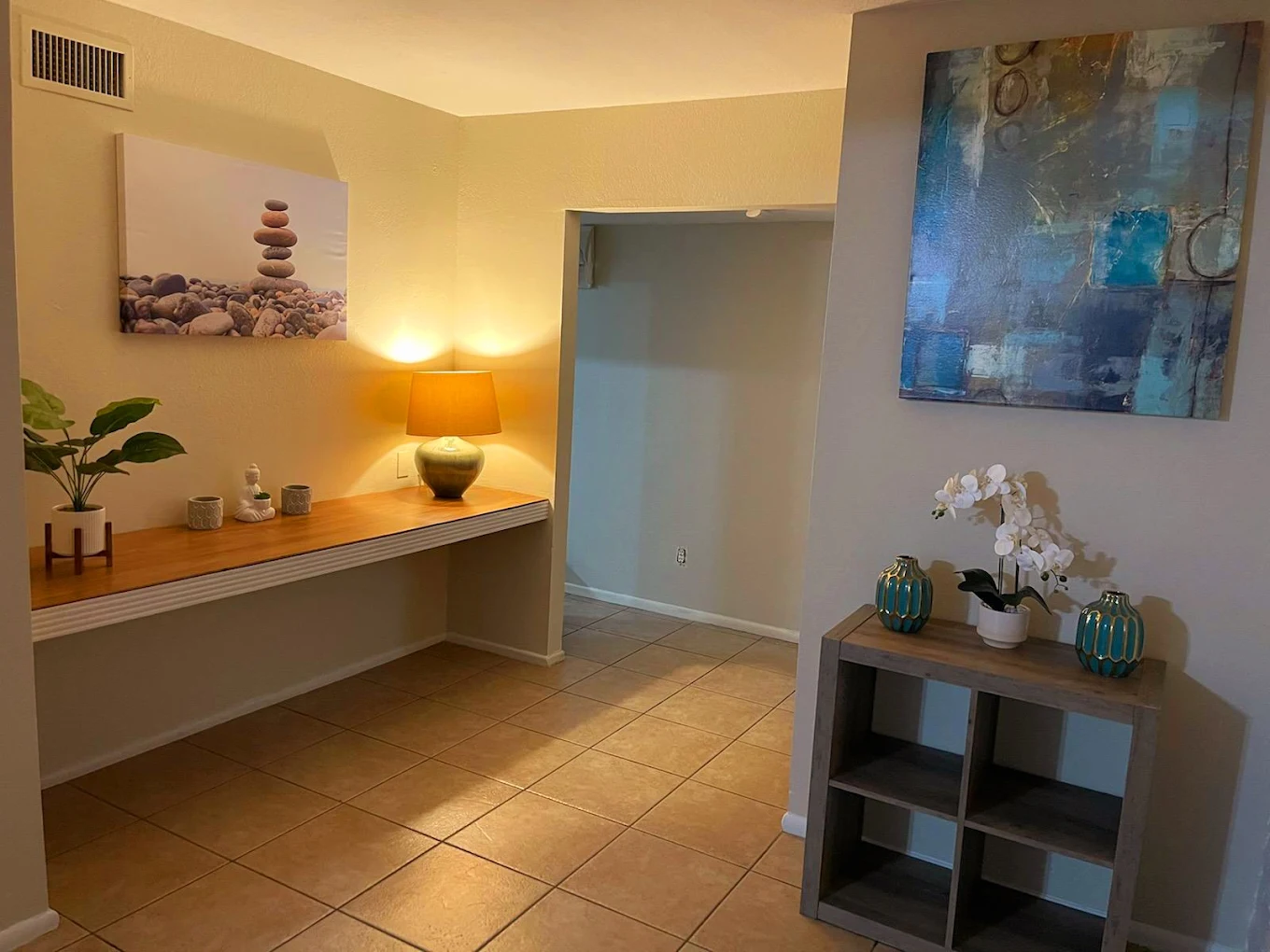 Interior view of a warmly lit hallway with a decorative shelf, potted plants, wall art, and a table lamp