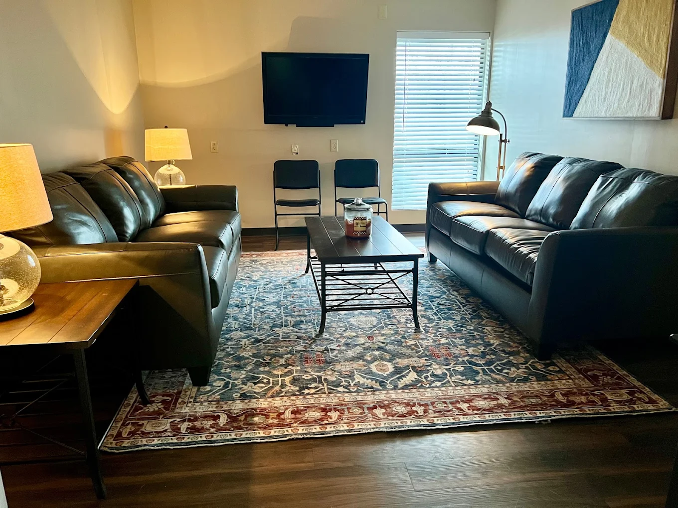 Cozy living room with two leather sofas, a coffee table, a wall-mounted TV, decorative rug, and natural light from window blinds