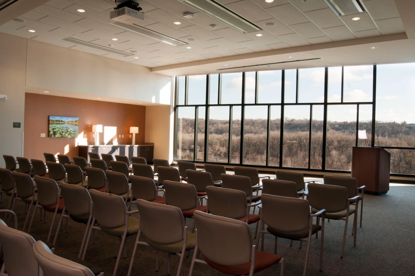 Large meeting room with rows of chairs, presentation podium, projector, and floor-to-ceiling windows overlooking a wooded landscape