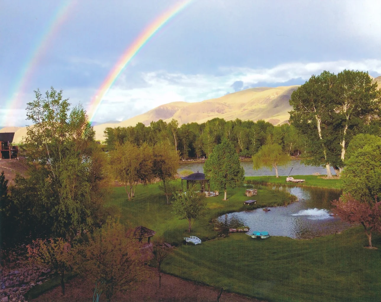 A vibrant double rainbow arcs over lush green lawns, a pond with a fountain, and distant rolling hills, highlighting the serene outdoor setting of Rainbows End Recovery Center in Challis.
