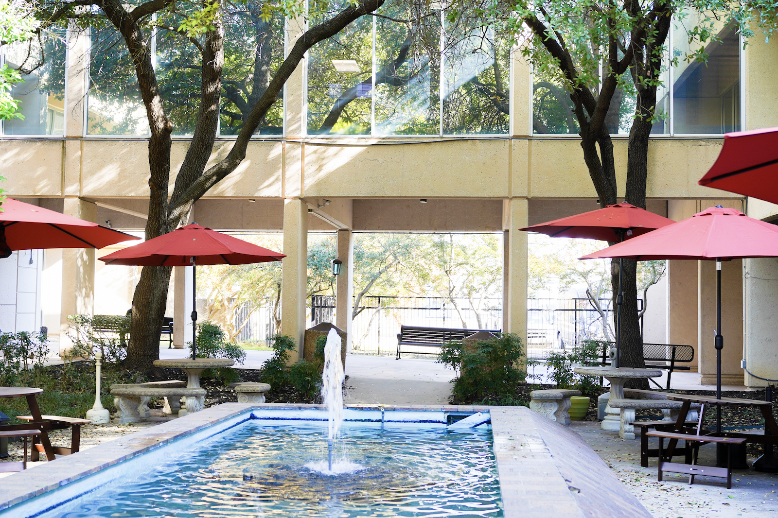 Shaded courtyard patio at Discovery Point Retreat Dallas with a central fountain, red umbrellas, picnic tables, benches, and mature trees