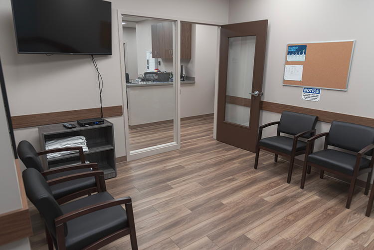 Small treatment and waiting area with black cushioned chairs, wall-mounted television, interior service window, bulletin board, and wood-look flooring
