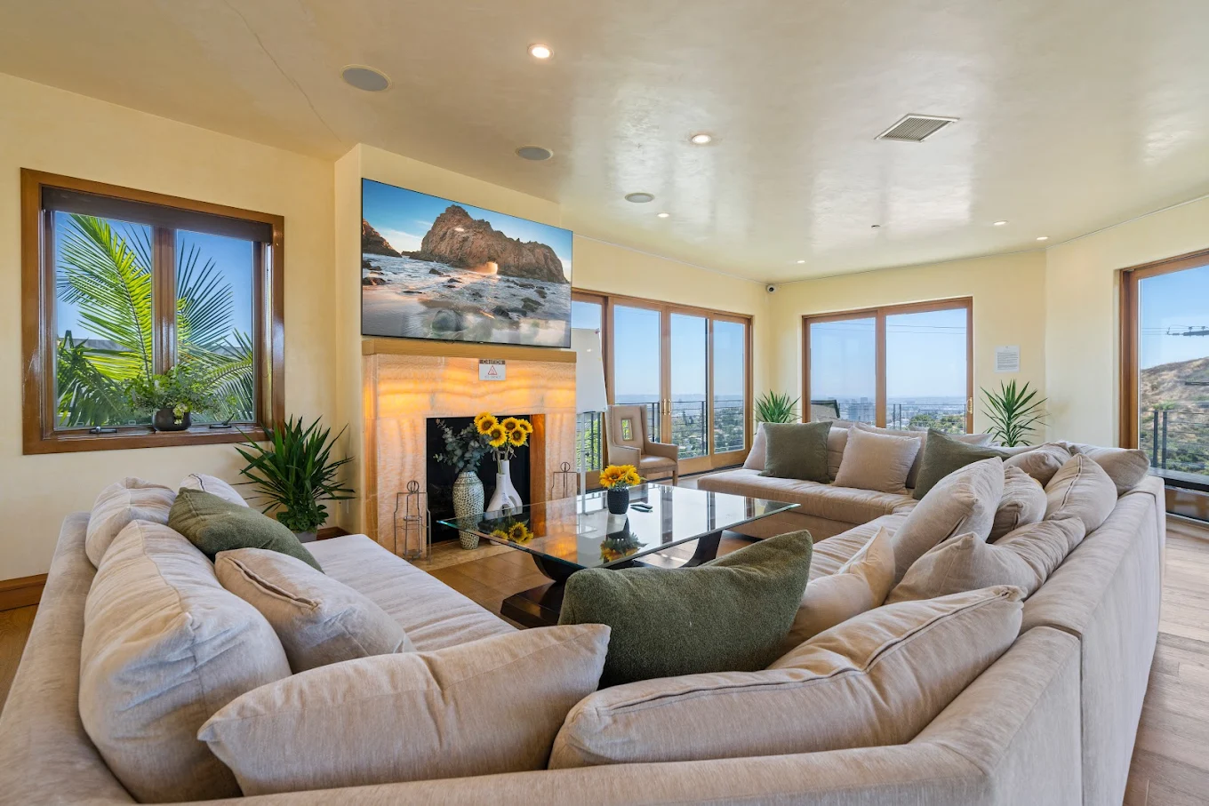 Bright living room with plush sectional sofas, a fireplace beneath a mounted TV, indoor plants, and large windows showcasing city and hillside views
