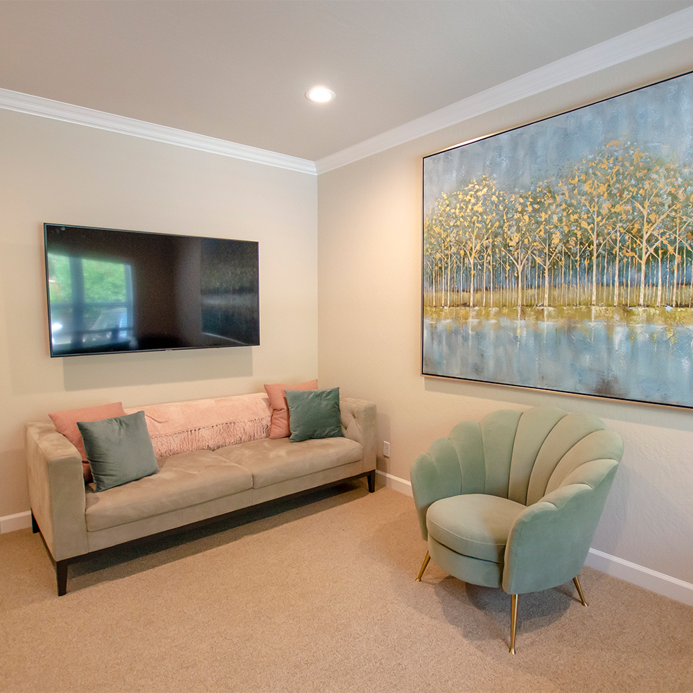  A cozy living space with a soft beige couch, pastel cushions, a stylish green armchair, a large nature-themed painting, and a mounted TV.