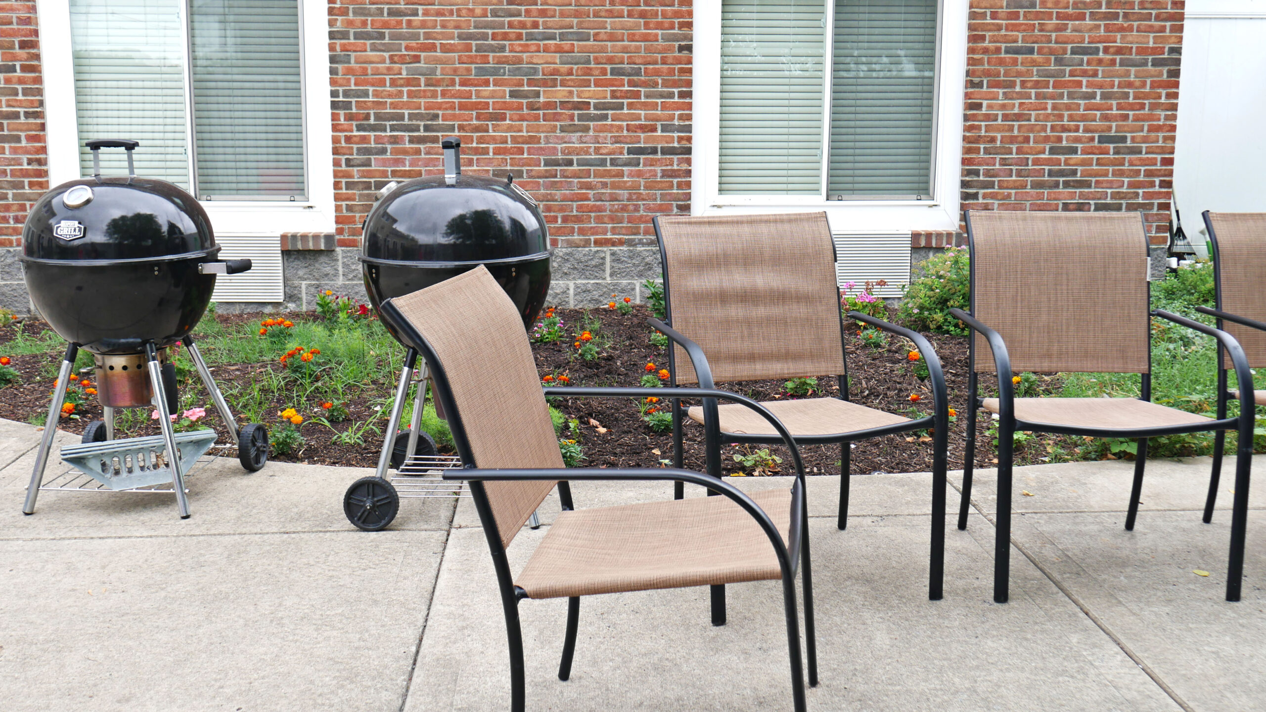 Outdoor patio with chairs and charcoal grills beside landscaped garden.