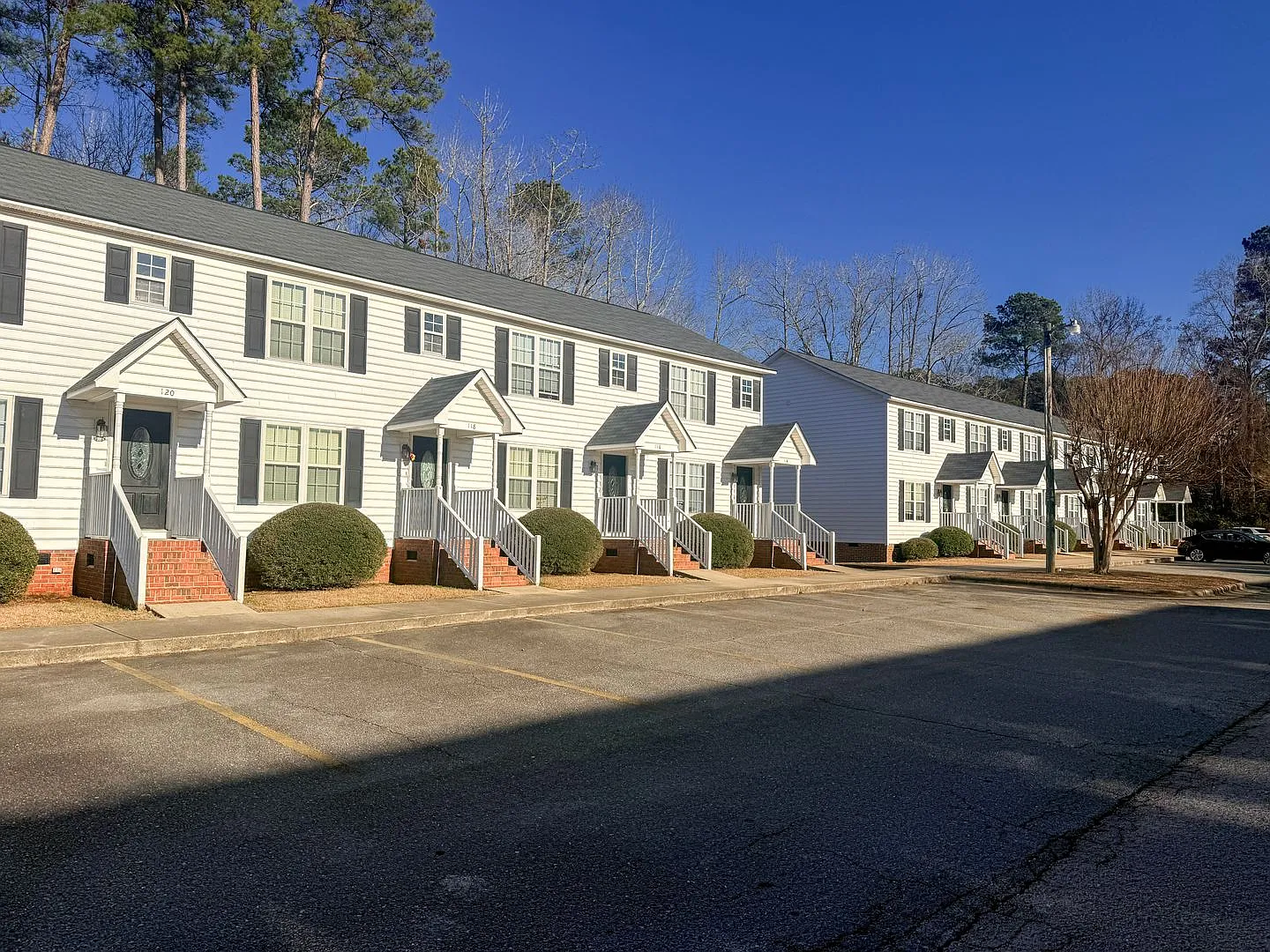 A row of white multi-unit townhouses with uniform porches, neatly trimmed bushes, and a spacious parking area in front.