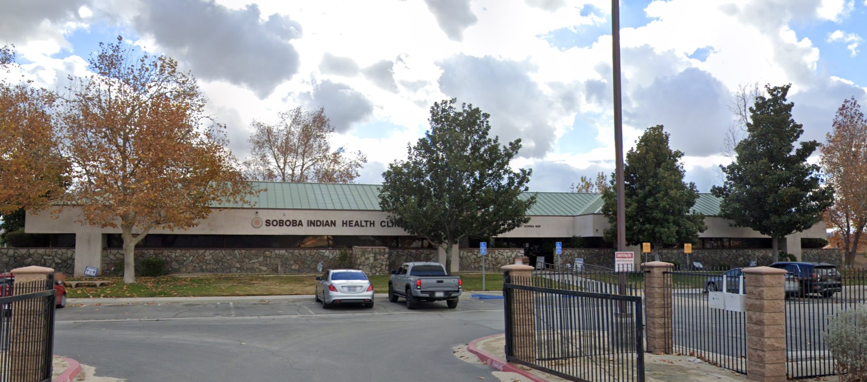 Entrance of the Soboba Indian Health Clinic, framed by seasonal trees and a gated parking area, offering easy access to visitors.