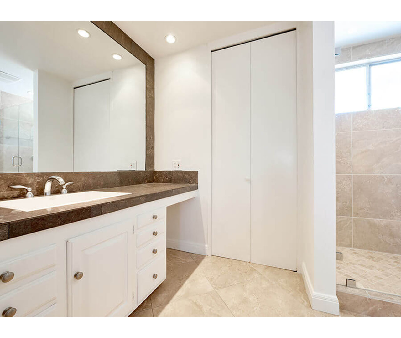A bright bathroom with a large mirror, white cabinets, and a tiled shower.