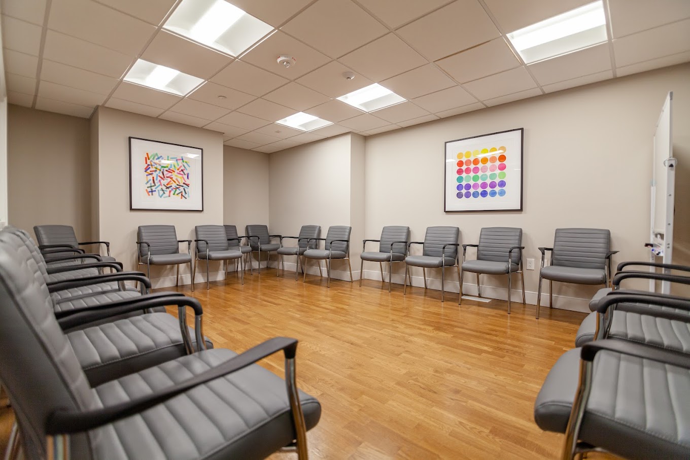  A well-lit group therapy room with rows of modern gray chairs arranged in a circle, light wooden flooring, and abstract artwork on the walls.