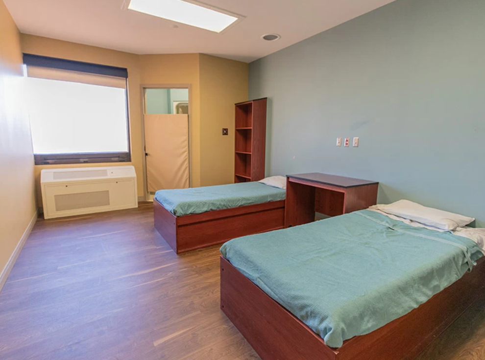 Two-bed patient room with teal bedding and wood furniture