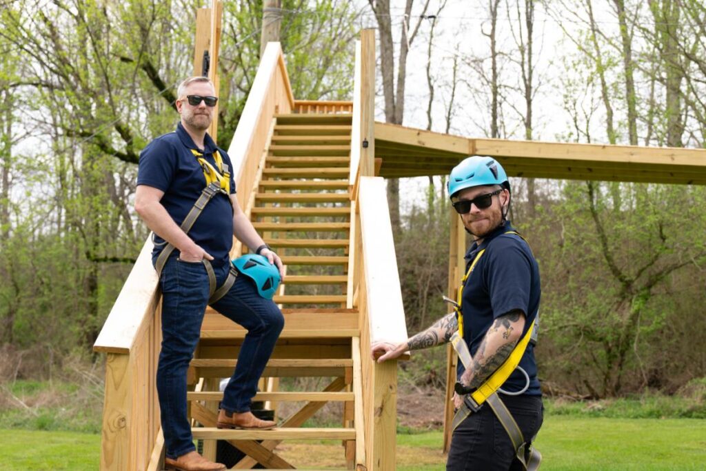 Two staff members in safety harnesses standing by the wooden stairs of a ropes course in a forested area