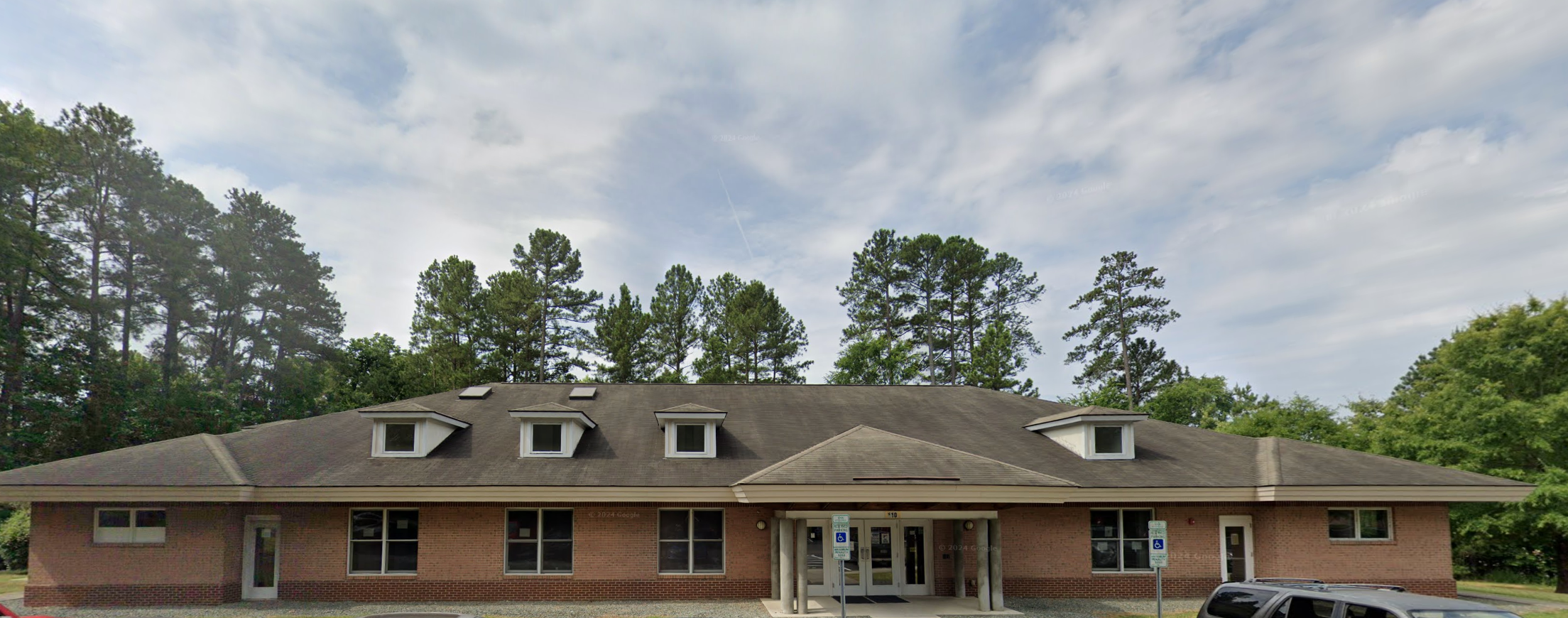 A front view of the Freedom House Recovery Center Men's Residential Program in Chapel Hill, showcasing a brick building with a sloped roof, surrounded by lush green trees.
