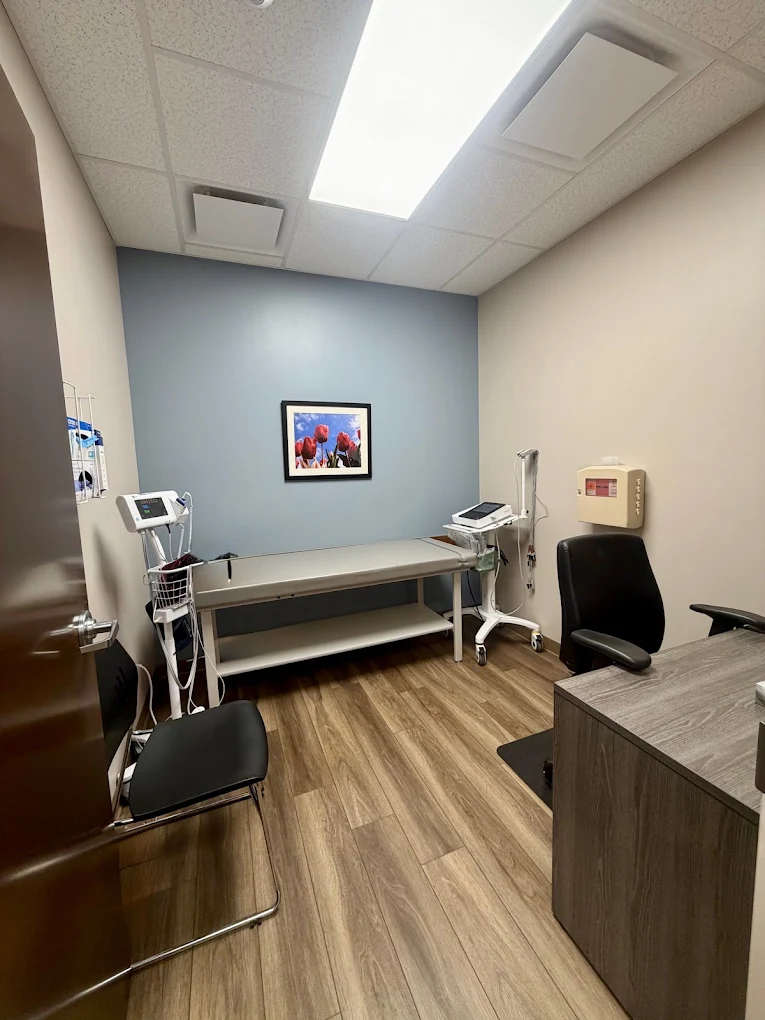 Clinical exam room with examination table, medical monitoring equipment, desk, chairs, blue accent wall, and framed artwork