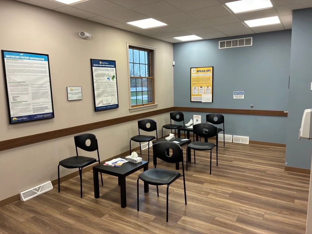 Treatment room with black chairs arranged for patients, small tables, informational posters, window, and wood-look flooring