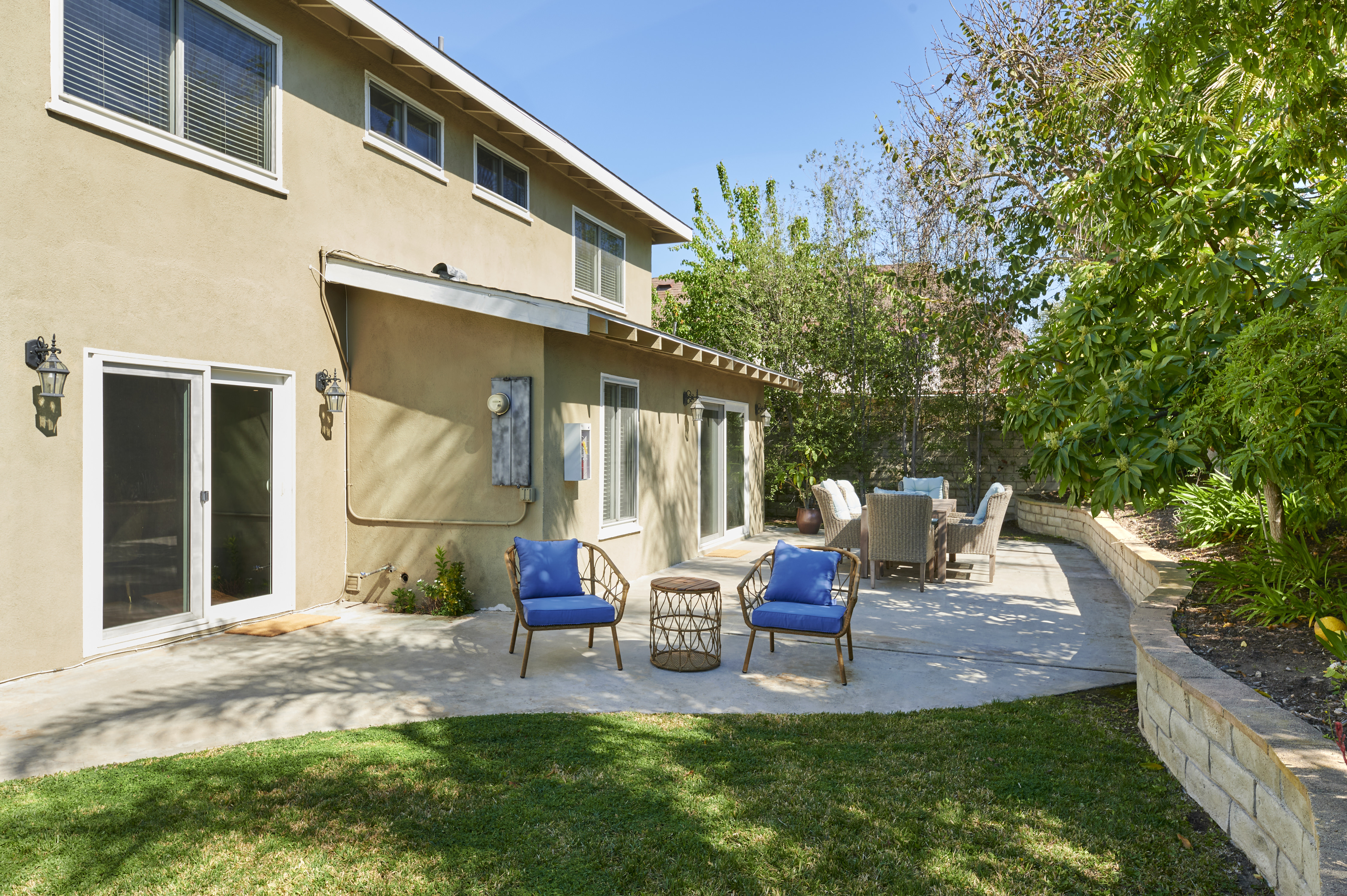 A sunny backyard patio with blue cushioned chairs and a cozy outdoor dining area, surrounded by lush greenery and trees, perfect for relaxation.
