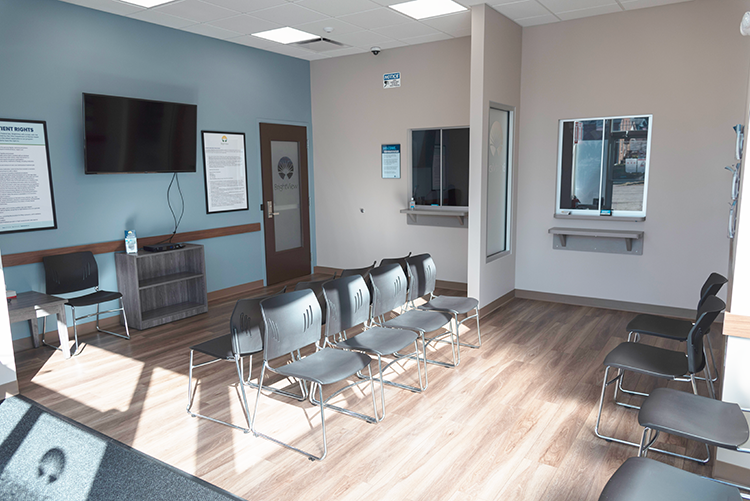 Bright waiting area at BrightView Health Mansfield with rows of black chairs, service windows, wall-mounted television, blue accent wall, and wood-look flooring
