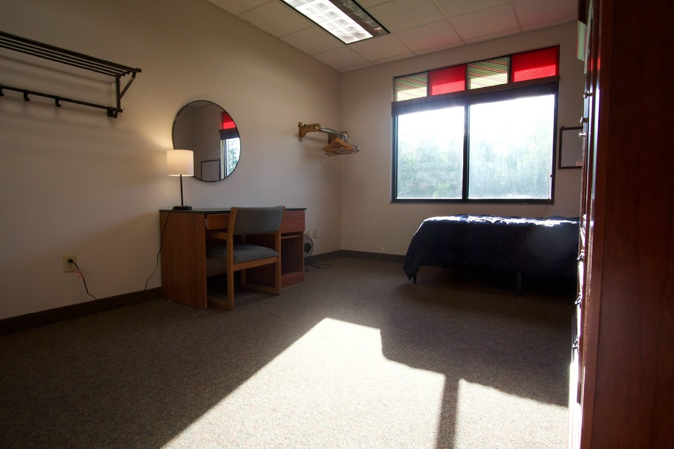 A bedroom with a desk, chair, mirror, and a single bed by the window