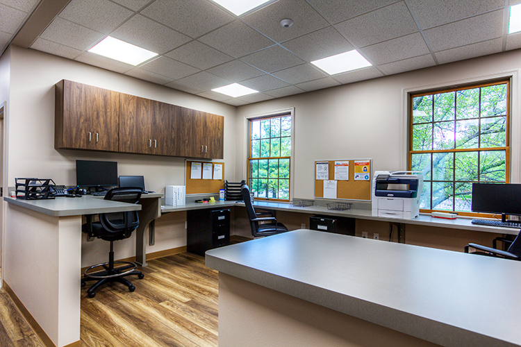 Staff work station with wraparound counters, computers, office chairs, printer, wood cabinets, and large windows