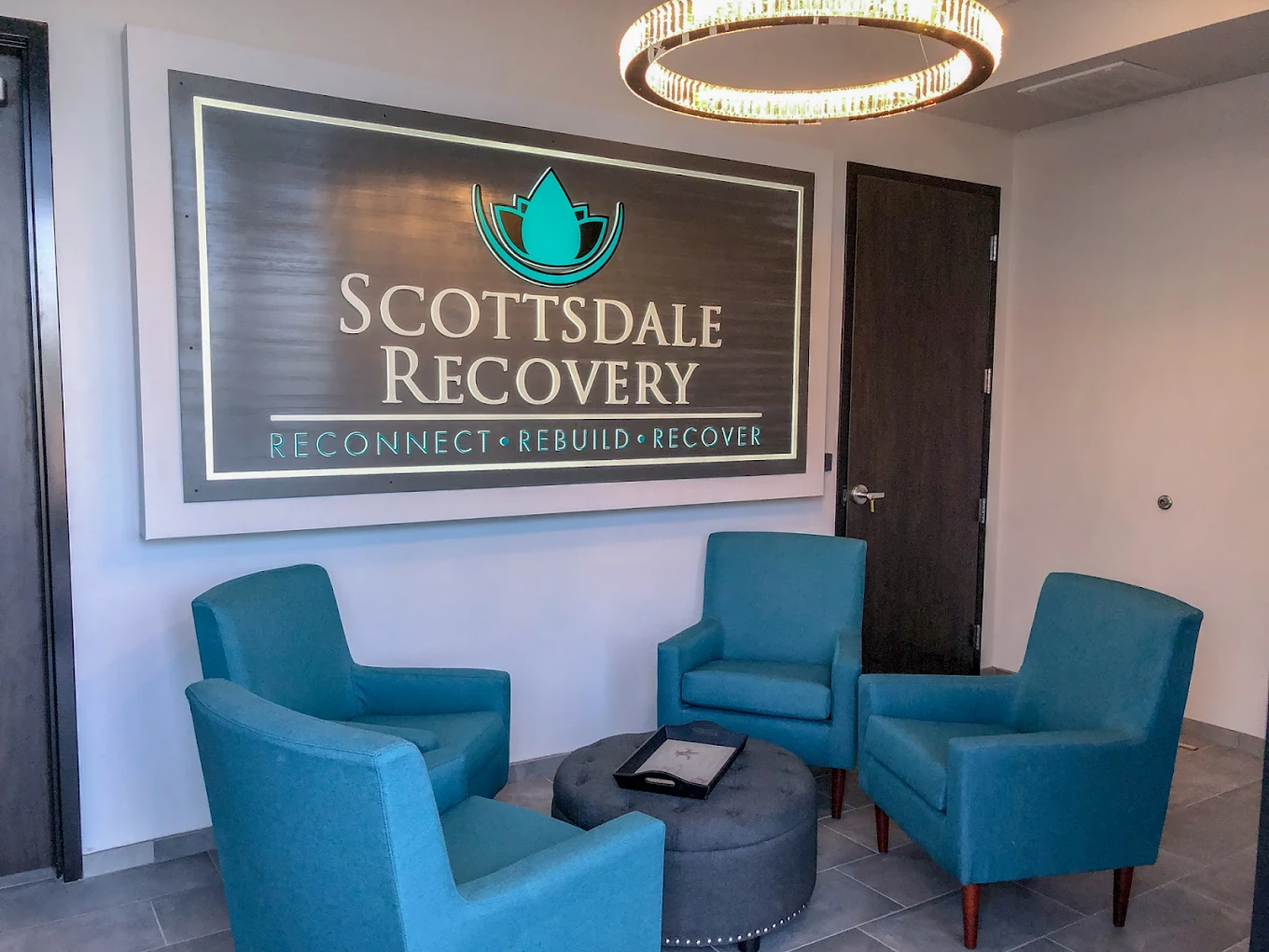 A modern lobby with a large Scottsdale Recovery sign on the wall, four teal armchairs arranged around a circular ottoman, and a contemporary chandelier overhead