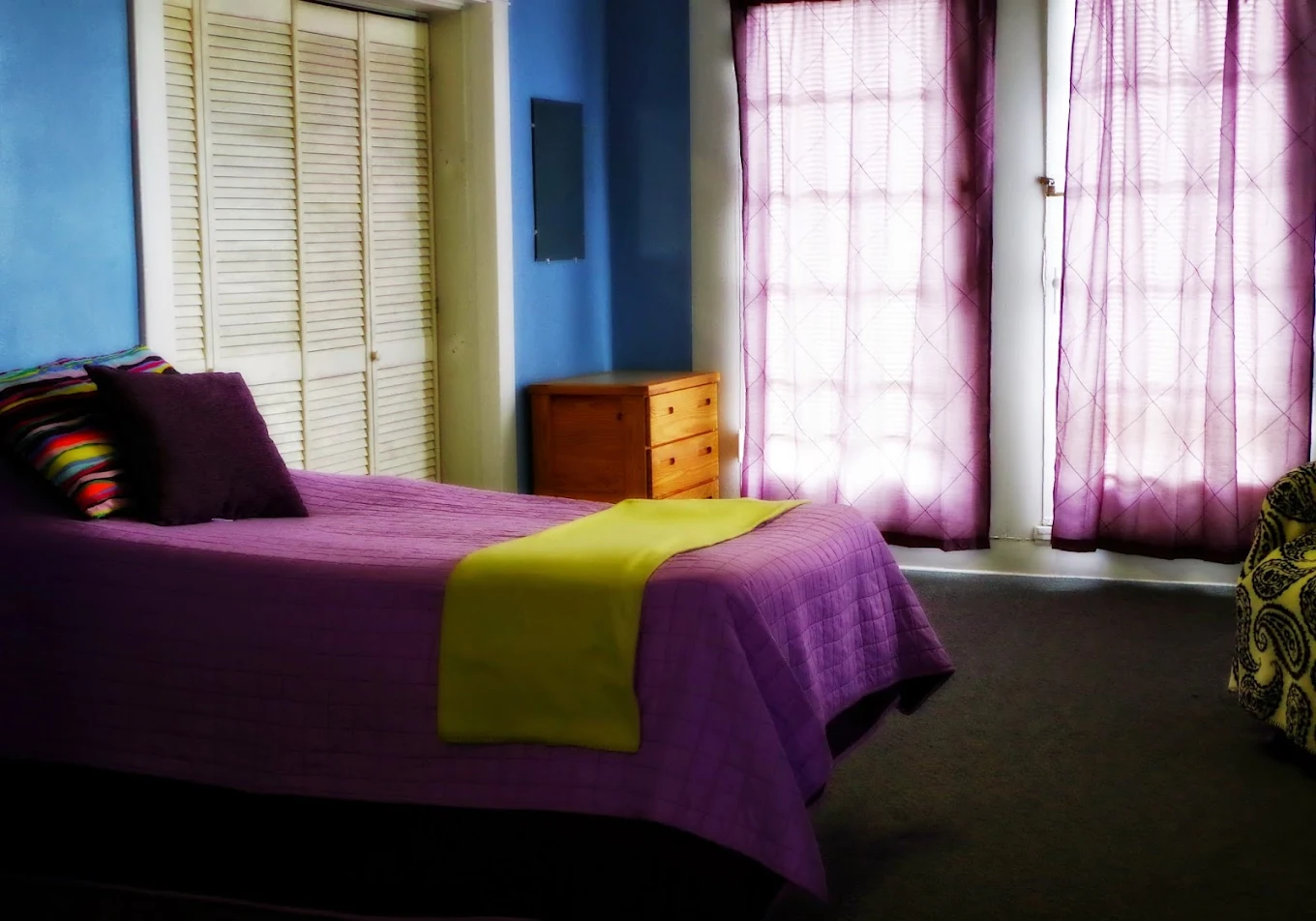 Rehab bedroom with purple bedding, bright curtains, and dresser