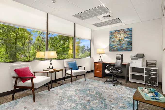 Bright office with chairs, desk, and large windows facing trees