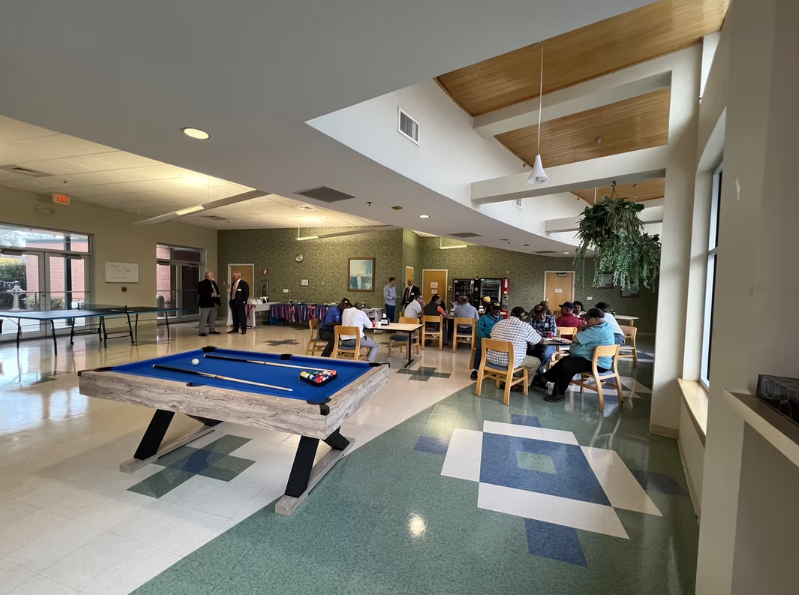 Common room with a pool table, people seated at tables, vending machines, and a ping-pong table in the background