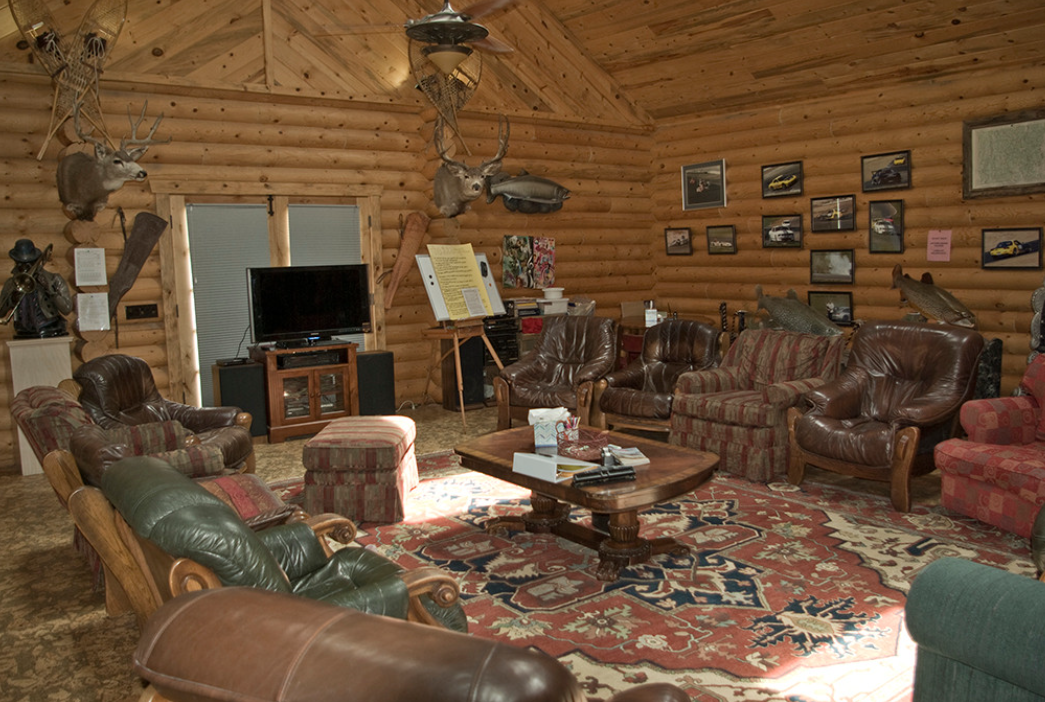 A spacious log-cabin style living room with leather armchairs and couches, mounted wildlife décor, a large rug, and warm wooden walls, creating a rustic and inviting atmosphere.
