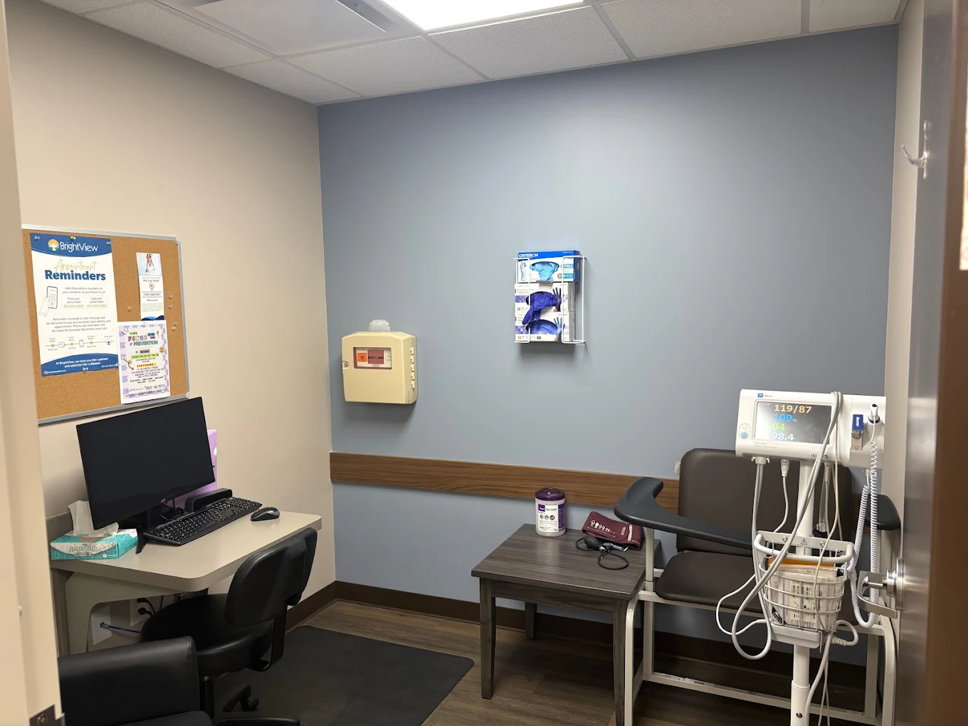 Clinical room at BrightView Health Marietta with computer workstation, patient chair, blood pressure and vital-sign monitoring equipment, medical supplies, and blue accent wall