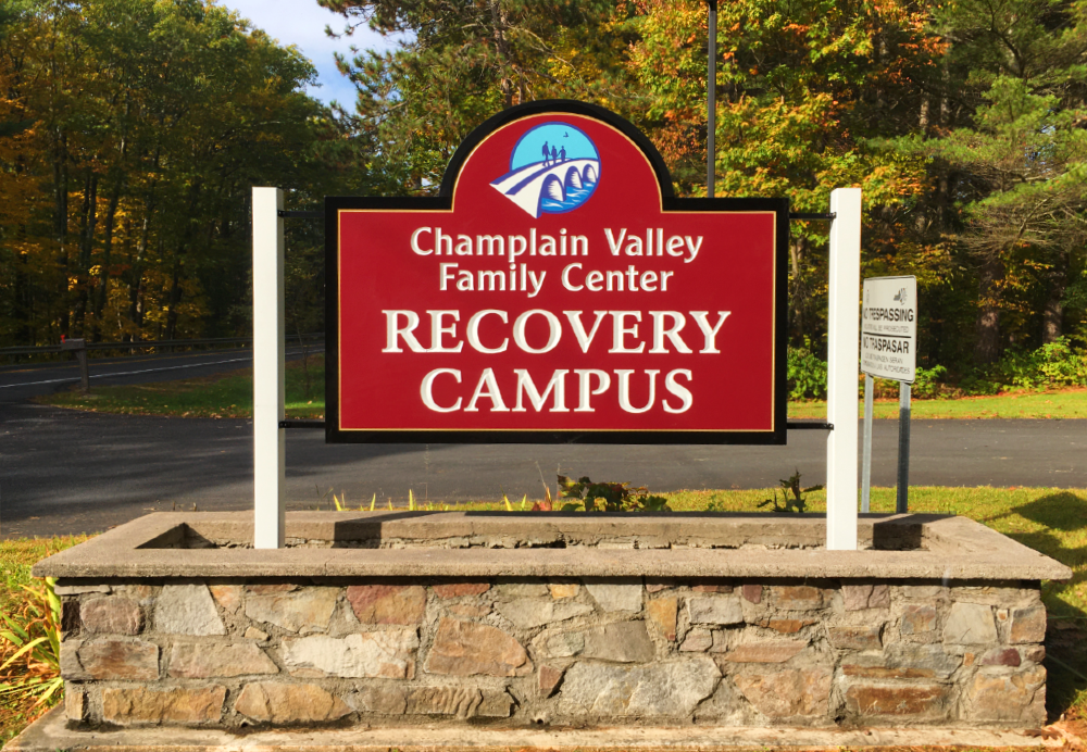 Red and white sign for Champlain Valley Family Center Recovery Campus displayed on a stone base near a wooded road