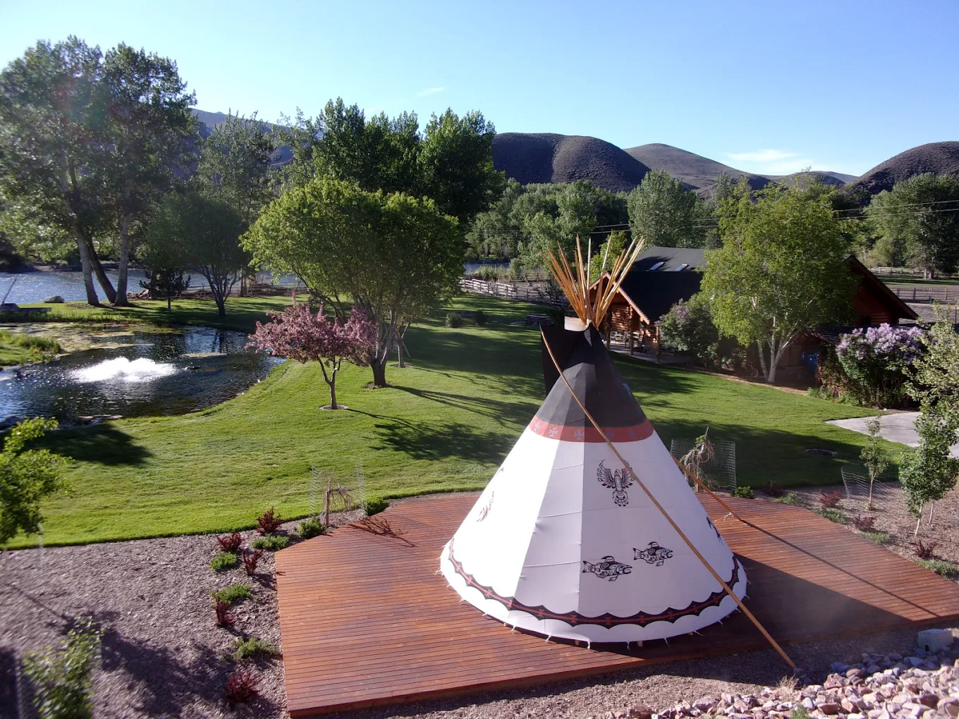 A large decorative teepee stands on a wooden platform surrounded by manicured lawns, trees, and a nearby pond with a fountain, creating a serene cultural and natural space.
